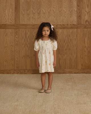 Rylee & Cru Brea Dress - Nutcracker
Young girl in a white dress with floral patterns standing against a wooden paneled wall.