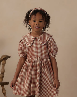 Rylee & Cru Camille Dress - Brick Check
A young girl with curly hair wearing a brown and beige gingham dress with a Peter Pan collar and puff sleeves, standing against a plain beige background.