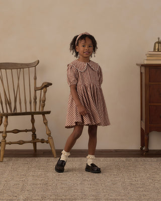 Rylee & Cru Camille Dress - Brick Check
A young girl stands in a vintage-style room wearing a short-sleeved gingham dress with a Peter Pan collar in a brown and white check pattern. She is styled with a matching headband, white ruffled socks, and black leather shoes.