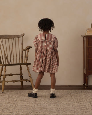 Rylee & Cru Camille Dress - Brick Check
A child stands with their back to the camera wearing a brown and white gingham dress, white socks, and black shoes in a vintage-style room with a wooden chair and cabinet.