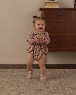 Rylee & Cru Cora Romper - Pink Floral
A toddler girl wearing a long-sleeve floral romper and pink booties is standing and smiling in a room with a wooden dresser and beige wall.