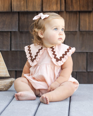 Rylee & Cru Cove Romper - Blush
Baby in a pink outfit with brown trim sitting on a wooden deck.