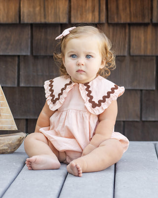 Rylee & Cru Cove Romper - Blush
Young child wearing a pink dress with a lace collar sitting on a wooden surface.