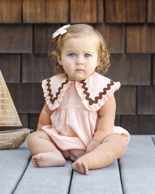 Rylee & Cru Cove Romper - Blush
Young child wearing a pink dress with brown trim sitting on a wooden surface.