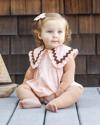 Rylee & Cru Cove Romper - BlushBaby in a pink dress with brown trim sitting on a wooden deck.
