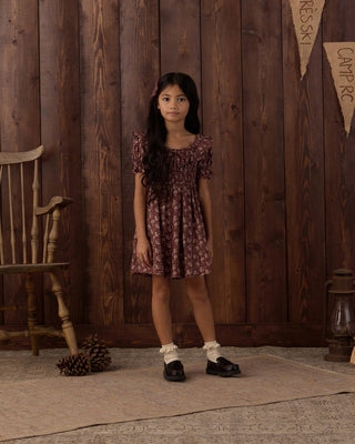 Rylee & Cru Dillon Dress - Mulberry Iris
A young girl stands in front of a rustic dark wood plank wall wearing a short-sleeved smocked dress with a dark red and brown floral pattern, white socks, and black shoes.