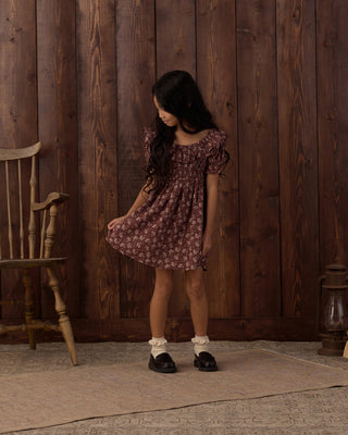 Rylee & Cru Dillon Dress - Mulberry Iris
A young girl with long dark hair stands in front of a rustic dark wood plank wall, wearing a short puff-sleeve dress with a dusty rose and white floral pattern. She is styled with white socks and black shoes, standing on a textured beige rug next to a wooden chair.