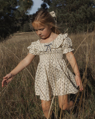 Rylee & Cru Dillon Dress - Vintage Fleur
A young girl wearing a cream-colored short-sleeve dress with a small floral print and a blue bow at the neckline, walking through a dry grassy field outdoors.