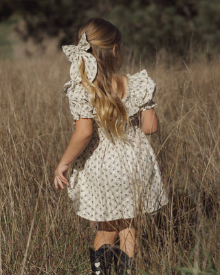 Rylee & Cru Dillon Dress - Vintage Fleur
A young girl with long blonde hair is seen from behind, wearing a cream-colored dress with a small floral pattern and a large matching hair bow, standing in a field of dry grass.