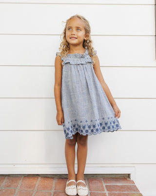 Rylee & Cru Lennon Dress - Marine Gingham
Young girl wearing a blue checkered dress standing against a white wooden wall.