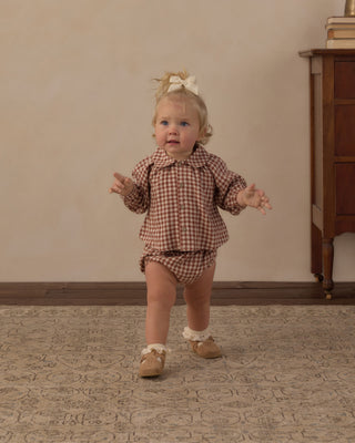 Rylee & Cru Nellie Set - Brick Check
A toddler girl wearing a brown and beige gingham long-sleeve romper and tan shoes, standing on a patterned rug in a studio setting.