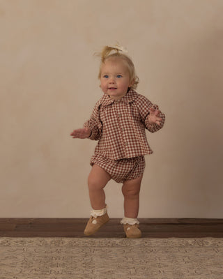 Rylee & Cru Nellie Set - Brick Check
A toddler girl wearing a matching brown and beige gingham two-piece outfit with a collared long-sleeve top and shorts, standing on a patterned rug against a neutral beige studio backdrop.