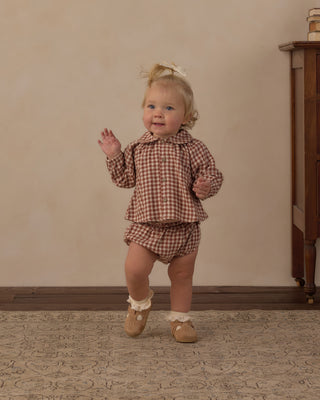 Rylee & Cru Nellie Set - Brick Check
A toddler girl wearing a matching brown and beige gingham long-sleeve shirt and shorts set, walking indoors on a patterned rug against a beige wall.