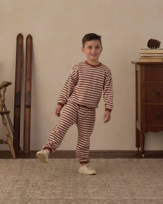 Rylee & Cru Relaxed Set - Brick Stripe
A young boy wearing a matching two-piece pajama set with horizontal rust red and cream stripes, standing in a room with a beige wall, wooden furniture, and vintage skis in the background.