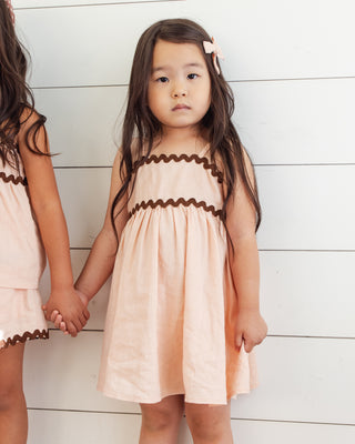 Rylee & Cru Ric Rac Dress - Blush
Two young girls wearing matching peach dresses with brown trim against a white wooden background.