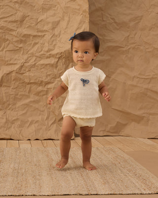 Rylee & Cru Scallop Knit Baby Set - Natural
Baby in a white outfit with butterfly designs standing on a textured brown floor.
