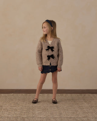 Rylee & Cru Scallop Mini Skirt - Dark Indigo
A young girl stands in a studio wearing a beige knit cardigan with two large black bow details, a dark navy mini skirt, and brown flats, set against a textured beige wall and patterned rug.