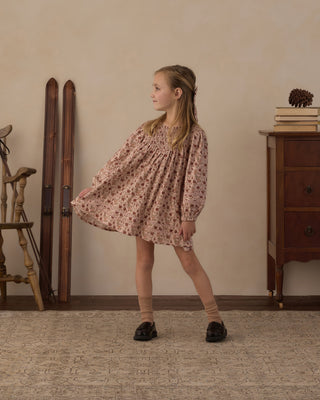 Rylee & Cru Selene Dress - Pink Floral
A young girl wearing a long-sleeved, smocked-bust mini dress with a dusty rose and brown floral pattern, standing in a vintage room with wooden furniture and skis in the background.