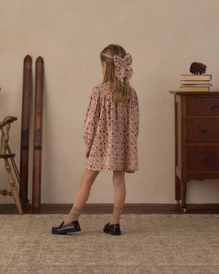 Rylee & Cru Selene Dress - Pink Floral 
A young girl is shown from behind wearing a dusty pink floral long-sleeve dress with a matching pink bow in her hair, styled with beige socks and black shoes in a vintage-style room.