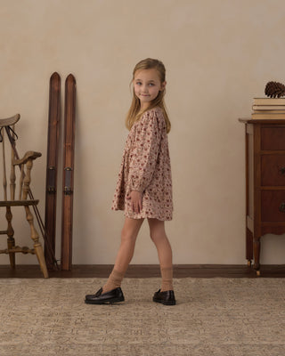 Rylee & Cru Selene Dress - Pink Floral
A young girl with blonde hair stands in a rustic room wearing a long-sleeved, knee-length pink dress with a small floral pattern, paired with beige socks and black loafers.