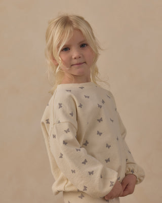 Rylee & Cru Spongy Knit Set - Butterfly Ditsy
A young blonde girl wearing a cream-colored long-sleeve sweatshirt with a small gray butterfly pattern, photographed against a warm beige studio background.