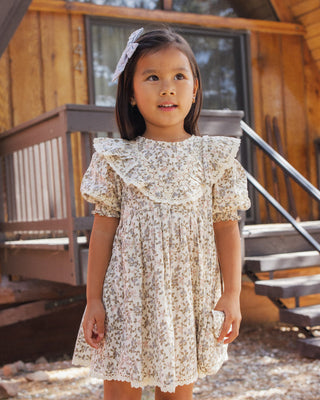 Rylee & Cru Winnie Dress - Winter Garden
A young girl wearing a cream-colored short-sleeve dress with a delicate floral print and a large ruffled collar, standing outdoors in front of a rustic wooden cabin.