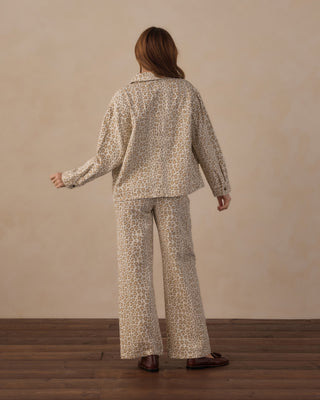 Rylee & Cru Women's Claire Jacket - Leopard
A woman stands with her back to the camera wearing a matching beige and brown animal-print pajama set, consisting of a long-sleeve button-up top and wide-leg pants, paired with brown leather loafers, against a neutral beige wall and wooden floor.