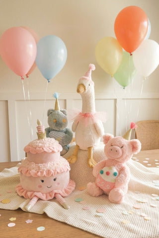 Mon Ami Birthday Goose
A soft pastel baby-themed party scene with plush toys and balloons: a pink ruffled cake plush, a white goose plush in a pink tutu, a pink pig plush, and a gray bear plush wearing a party hat, arranged on a cream blanket with confetti. Colorful balloons in pink, blue, orange, yellow, green, and white float in the background against a light wall.