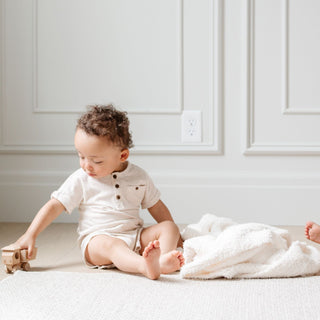 Saranoni Bamboni® Receiving Blanket - Ivory
 Child sitting on a white blanket playing with a toy in a room with white walls.