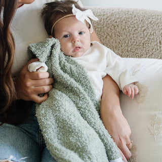 Saranoni Bamboni® Receiving Blanket - Sage
Baby wrapped in a green blanket with a white headband, held by an adult on a beige couch.