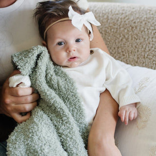 Saranoni Bamboni® Receiving Blanket - Sage
Baby wrapped in a green blanket with a white headband, sitting on a beige surface.