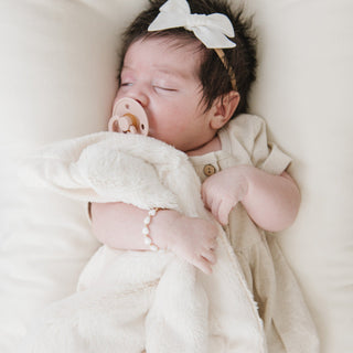Saranoni Lush Mini Blanket - Natural
Newborn baby sleeping peacefully with a pacifier and white bow headband, wrapped in a soft blanket.