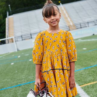 Pink Chicken Girls Maribelle Dress - Flying Footballs Young girl in a yellow dress with a pattern, standing on a sports field.