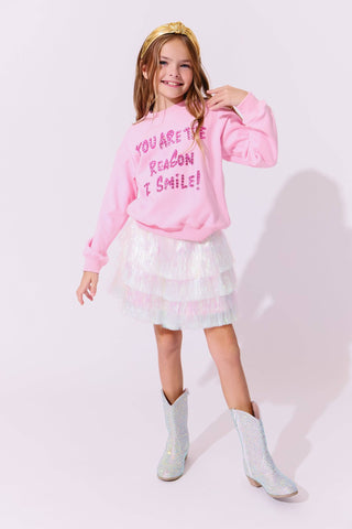 Lola and The Boys Snow Shimmer Tinsel Party Skirt
Young girl wearing a pink sweatshirt with text, white dress, and light blue boots on a white background
