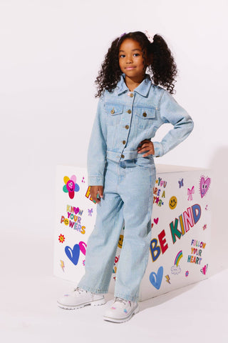 Lola and The Boys Sparkle Crystal Denim Jeans
A young girl with curly hair stands in a studio wearing a light blue denim jumpsuit and white sneakers, posing next to a white box decorated with colorful 'BE KIND' and heart graphics.