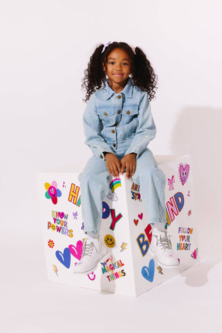 Lola and The Boys Sparkle Crystal Denim Jeans
A young girl with curly hair in pigtails sits on a white decorative box covered in colorful motivational stickers. She is wearing a light blue denim jacket and matching jeans with white sneakers, set against a plain white studio background.