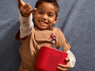Tonies Character - MARVEL Spider-Man
Child wearing a brown outfit with a superhero figure, holding a red toy box against a textured blue background.