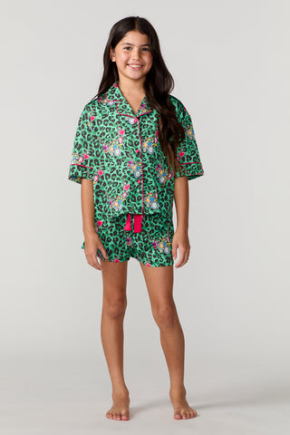 Sunshine Society Kids Silky Short Sleeve Short Set - Gilded Leopard
A young girl stands barefoot against a light gray studio background wearing a short-sleeve button-front pajama shirt and matching shorts set. The pajamas have a green leopard print base with pink and white floral motifs and a bright pink waist tie.