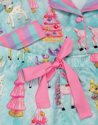 Sunshine Society Kids Silky Short Sleeve Short Set - Prancer
A close-up of a children's pajama set featuring a light blue and pink holiday print with deer, Christmas trees, and snowflakes, complete with a large pink bow at the waist.
