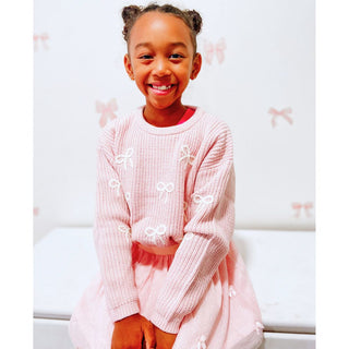 Sweet Wink All Over Bows Knit Sweater
Young girl wearing a pink outfit with white embellishments, sitting on a white surface.