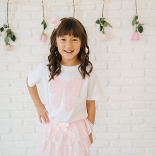 Sweet Wink Bow Lace Tiered Tutu
Young girl wearing a white t-shirt with pink heart design and pink skirt against a white brick wall.
