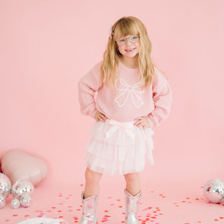 Sweet Wink Bow Lace Tiered Tutu
Young girl in a pink sweater and white skirt standing on a pink background with decorative elements.