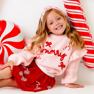 Sweet Wink Candy Cane Christmas Gemstone Headband - Kids
Sweet Wink Coquette Merry Christmas Knit Sweater
Young girl in a pink sweater with 'Merry' text, red skirt, and pink headband surrounded by candy-themed decorations.