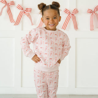 Sweet Wink Candy Cane Lane Christmas Sweatshirt & Flare Legging Set
Child wearing a pink pajama set with red bows against a white background with red bow decorations.
