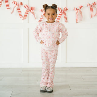 Sweet Wink Candy Cane Lane Christmas Sweatshirt & Flare Legging Set
Child wearing pink pajamas with red patterns in a room with decorative bows on the wall.