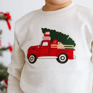 Sweet Wink Christmas Truck Patch Sweatshirt - Natural 
Beige sweatshirt with a red truck and Christmas tree design on a blurred festive background