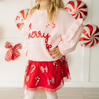 Sweet Wink Coquette Merry Christmas Knit Sweater
Child wearing a pink sweater with red text and a red skirt with candy cane patterns, standing in front of candy-themed decorations.