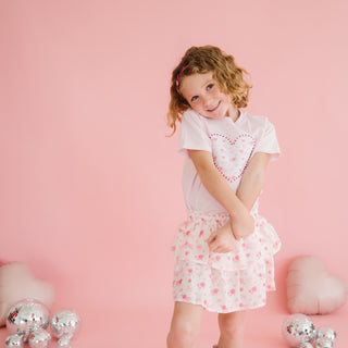 Sweet Wink Dainty Rose Skort
Young girl in a white dress with pink heart patterns on a pink background