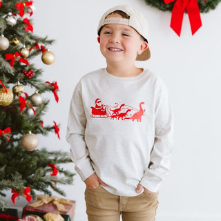 Sweet Wink Dino Christmas Long Sleeve Shirt - Natural
Child wearing a white sweatshirt with red graphics in front of a decorated Christmas tree.