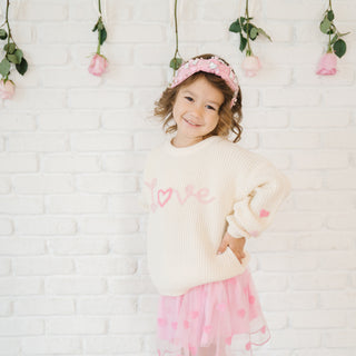 Sweet Wink Ditsy Love Valentine's Day Knit Sweater Young girl wearing a cream sweater with pink hearts and a pink skirt, standing against a white brick wall with floral decorations.